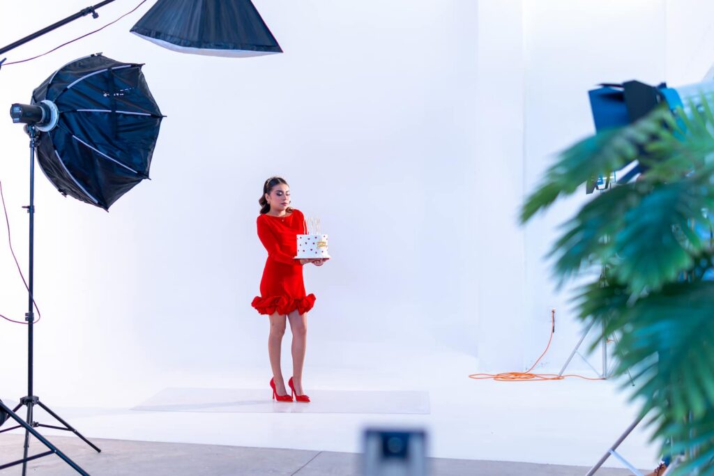 A behind-the-scenes view of a commercial photography studio showing a woman in a red ruffled dress and red heels holding a polka-dot gift box on a white seamless backdrop, surrounded by professional lighting equipment and softboxes.