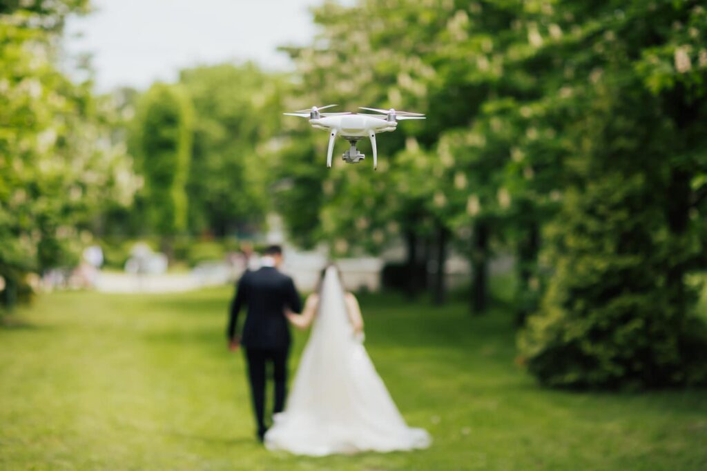A white drone hovering in focus in the foreground, filming a blurred bride and groom walking hand-in-hand through a grassy park in the background.